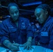 Two U.S. Navy personnel in blue utility uniforms are working together at a workstation aboard what appears to be a naval vessel. The individual on the left, identified by their name tape as 'PASCASIO,' is writing on a document while the other person observes. The environment is illuminated by blue lighting, typical of a ship's combat information center or navigation space.