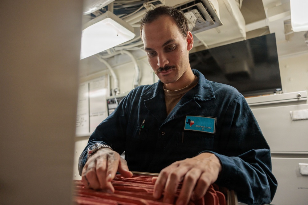 A male service member wearing a dark blue uniform/coverall is working with what appears to be a red filing or storage tray in an interior space consistent with a naval vessel. His name tag reads 'Donovan Lopez' and features a Texas state outline graphic. He appears focused on organizing or retrieving items from the red tray.