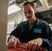 A male service member wearing a dark blue uniform/coverall is working with what appears to be a red filing or storage tray in an interior space consistent with a naval vessel. His name tag reads 'Donovan Lopez' and features a Texas state outline graphic. He appears focused on organizing or retrieving items from the red tray.