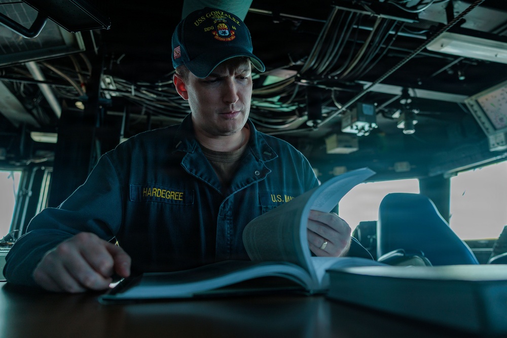 A U.S. Navy sailor named Hardegree is seated at a workstation aboard a naval vessel, reviewing documents or a manual. He is wearing a navy blue uniform with 'U.S. NAVY' and 'HARDEGREE' name tape visible, along with a USS Gonzalez (DDG) ship's cap. The setting appears to be the bridge or a command area of the ship, with navigation equipment and overhead cabling visible in the background.