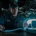 A U.S. Navy sailor named Hardegree is seated at a workstation aboard a naval vessel, reviewing documents or a manual. He is wearing a navy blue uniform with 'U.S. NAVY' and 'HARDEGREE' name tape visible, along with a USS Gonzalez (DDG) ship's cap. The setting appears to be the bridge or a command area of the ship, with navigation equipment and overhead cabling visible in the background.