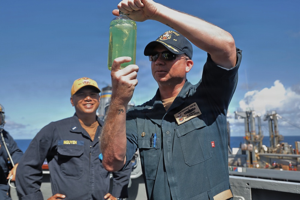 A U.S. Navy officer, identified by his name tag as a Commanding Officer aboard USS Farragut (DDG 99), holds up a glass bottle containing a green liquid for inspection on the ship's deck. A second sailor named Nguyen, wearing a yellow cap, stands in the background smiling. The scene appears to take place during an at-sea operation with another vessel visible in the background.