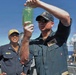A U.S. Navy officer, identified by his name tag as a Commanding Officer aboard USS Farragut (DDG 99), holds up a glass bottle containing a green liquid for inspection on the ship's deck. A second sailor named Nguyen, wearing a yellow cap, stands in the background smiling. The scene appears to take place during an at-sea operation with another vessel visible in the background.