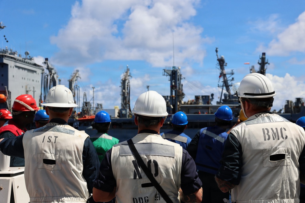 A group of naval or maritime personnel wearing hard hats and safety vests are gathered on a ship deck, facing toward other vessels in what appears to be a naval port or at-sea replenishment operation. Several individuals have markings on their vests including 'IST', 'BMC', and 'NO' along with 'DDG 49' visible on one vest. The scene suggests a briefing, muster, or observation of a ship-to-ship operation.