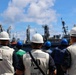 A group of naval or maritime personnel wearing hard hats and safety vests are gathered on a ship deck, facing toward other vessels in what appears to be a naval port or at-sea replenishment operation. Several individuals have markings on their vests including 'IST', 'BMC', and 'NO' along with 'DDG 49' visible on one vest. The scene suggests a briefing, muster, or observation of a ship-to-ship operation.