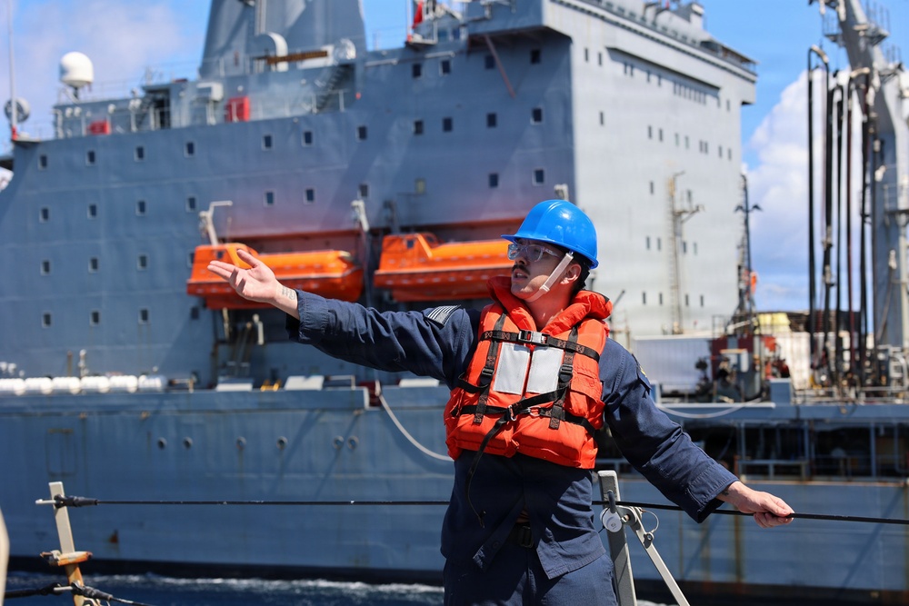 A naval or maritime crew member wearing a blue hard hat, orange life vest, and safety glasses is giving hand signals or directing operations on the deck of a vessel. In the background, a large gray military or auxiliary ship is visible alongside, with orange enclosed lifeboats mounted on its hull. The scene appears to depict an underway replenishment (UNREP) or ship-to-ship operation at sea.