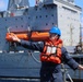 A naval or maritime crew member wearing a blue hard hat, orange life vest, and safety glasses is giving hand signals or directing operations on the deck of a vessel. In the background, a large gray military or auxiliary ship is visible alongside, with orange enclosed lifeboats mounted on its hull. The scene appears to depict an underway replenishment (UNREP) or ship-to-ship operation at sea.