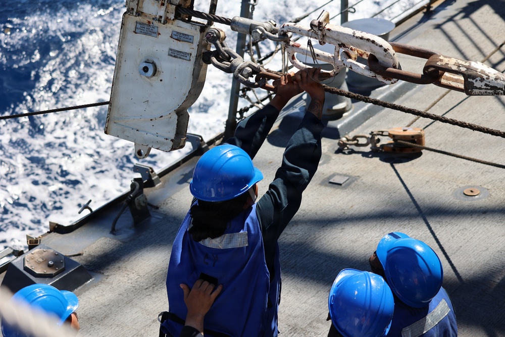 Several workers wearing blue hard hats and blue coveralls are performing maintenance or rigging work on the deck of a ship at sea. One worker is reaching upward to handle a chain and shackle assembly attached to overhead rigging equipment. The ocean is visible in the background, with white-capped waves indicating active sea conditions.