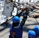 Several workers wearing blue hard hats and blue coveralls are performing maintenance or rigging work on the deck of a ship at sea. One worker is reaching upward to handle a chain and shackle assembly attached to overhead rigging equipment. The ocean is visible in the background, with white-capped waves indicating active sea conditions.
