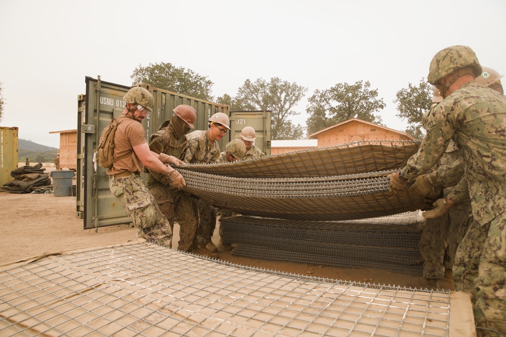 A group of military personnel in camouflage uniforms and hard hats are working together to handle large sections of wire mesh or reinforcement grid material. The scene appears to be a construction or engineering training exercise at a military installation, with shipping containers and partially constructed wooden structures visible in the background. The personnel appear to be Navy Seabees or similar military construction unit based on their uniforms and equipment.