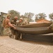 A group of military personnel in camouflage uniforms and hard hats are working together to handle large sections of wire mesh or reinforcement grid material. The scene appears to be a construction or engineering training exercise at a military installation, with shipping containers and partially constructed wooden structures visible in the background. The personnel appear to be Navy Seabees or similar military construction unit based on their uniforms and equipment.