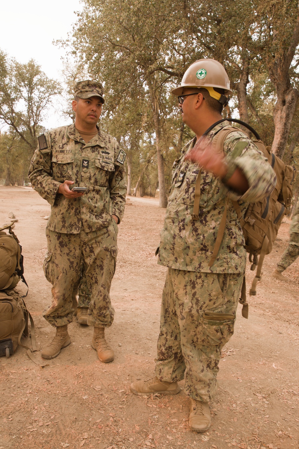 Two U.S. Navy Seabees in NWU Type III (AOR-2) camouflage uniforms are engaged in conversation in an outdoor wooded area with dry, sandy terrain. The sailor on the left, identified by the name tape 'FERRETTI' and 'U.S. NAVY' tape, is holding a mobile device. The sailor on the right is wearing a white hard hat with a green medical cross symbol and carrying a tactical backpack.