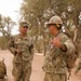 Two U.S. Navy Seabees in NWU Type III (AOR-2) camouflage uniforms are engaged in conversation in an outdoor wooded area with dry, sandy terrain. The sailor on the left, identified by the name tape 'FERRETTI' and 'U.S. NAVY' tape, is holding a mobile device. The sailor on the right is wearing a white hard hat with a green medical cross symbol and carrying a tactical backpack.
