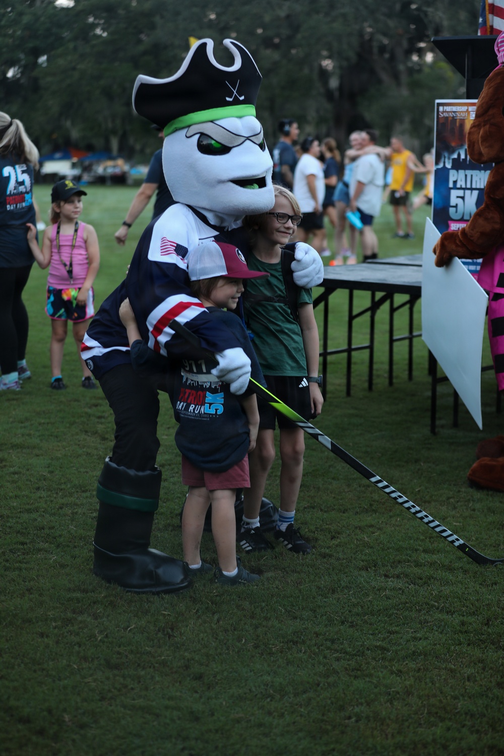 A sports mascot dressed as a pirate-themed hockey character poses with two children at an outdoor event. The mascot holds a Warrior-branded hockey stick and wears a jersey with an American flag patch. A banner in the background references a 'Patriot 5K' event, suggesting this is a community race or fundraiser.