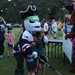 A sports mascot dressed as a pirate-themed hockey character poses with two children at an outdoor event. The mascot holds a Warrior-branded hockey stick and wears a jersey with an American flag patch. A banner in the background references a 'Patriot 5K' event, suggesting this is a community race or fundraiser.