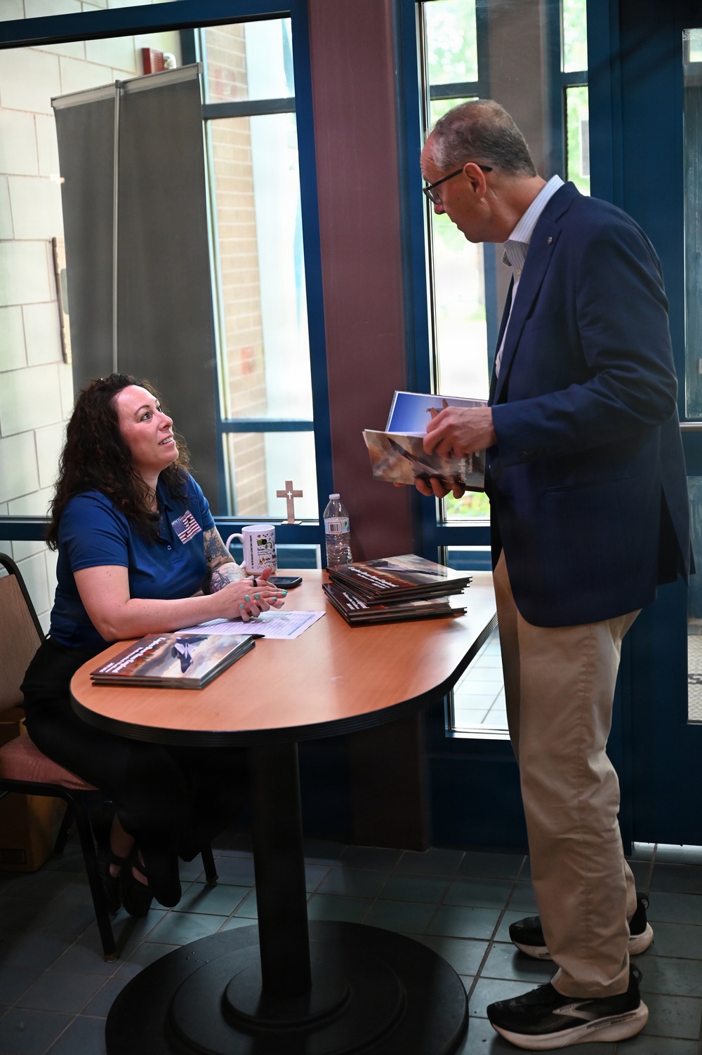 A woman seated at a round table is engaged in conversation with a standing older man who is holding printed materials or a booklet. The setting appears to be an indoor lobby or common area, possibly at a conference, fair, or informational event. The woman is wearing a blue polo shirt with an American flag patch and has visible tattoos on her arm.