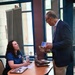 A woman seated at a round table is engaged in conversation with a standing older man who is holding printed materials or a booklet. The setting appears to be an indoor lobby or common area, possibly at a conference, fair, or informational event. The woman is wearing a blue polo shirt with an American flag patch and has visible tattoos on her arm.