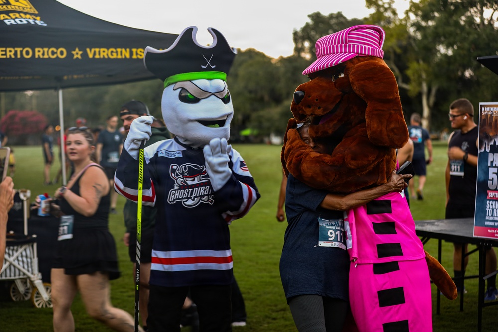 An outdoor event featuring two mascot characters — one wearing a 'Ghost Pirates' hockey jersey with a pirate hat and holding a hockey stick, and another dressed as a brown dog in pink outfit and striped hat. The event appears to be a patriot-themed run or charity event, indicated by race bibs and signage. An Army ROTC tent with 'Puerto Rico ★ Virgin Islands' text is visible in the background.