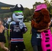 An outdoor event featuring two mascot characters — one wearing a 'Ghost Pirates' hockey jersey with a pirate hat and holding a hockey stick, and another dressed as a brown dog in pink outfit and striped hat. The event appears to be a patriot-themed run or charity event, indicated by race bibs and signage. An Army ROTC tent with 'Puerto Rico ★ Virgin Islands' text is visible in the background.