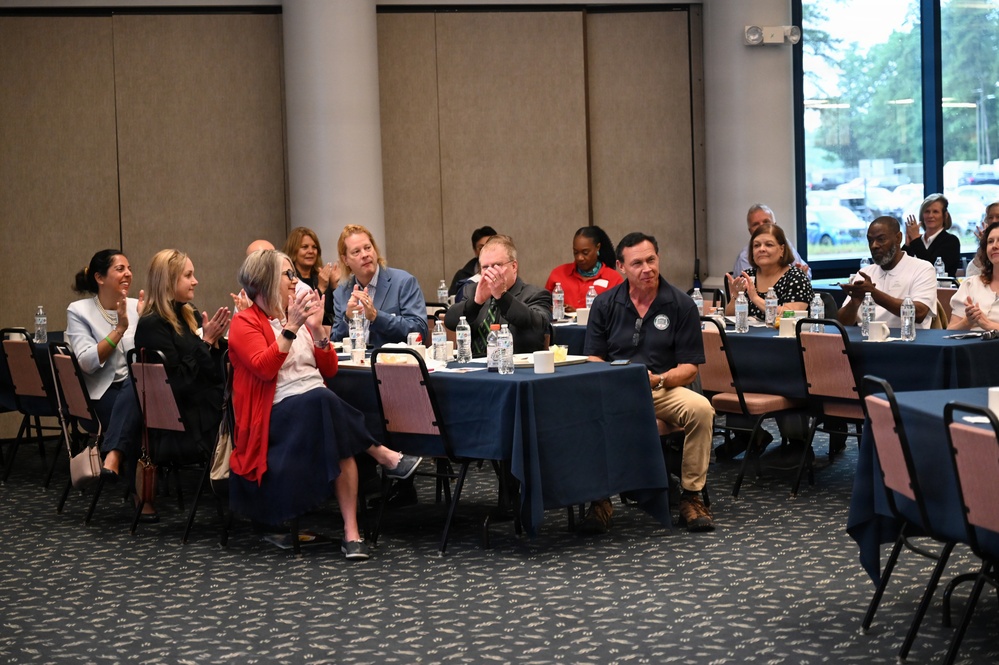A group of approximately 15-20 adults are seated at round tables covered with navy blue tablecloths in what appears to be a conference or banquet hall. Most attendees are clapping or applauding, suggesting they are responding to a presentation or announcement. The setting appears to be a professional or organizational meeting event.