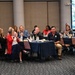 A group of approximately 15-20 adults are seated at round tables covered with navy blue tablecloths in what appears to be a conference or banquet hall. Most attendees are clapping or applauding, suggesting they are responding to a presentation or announcement. The setting appears to be a professional or organizational meeting event.