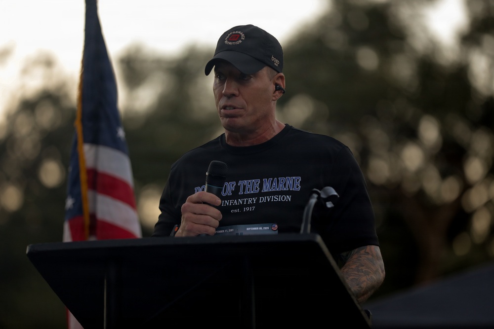 A middle-aged man stands at a podium speaking into a handheld microphone at what appears to be an outdoor commemorative event. He is wearing a black t-shirt referencing the 3rd Infantry Division ('Rock of the Marne, est. 1917') and a black baseball cap. An American flag is visible in the background to his left.