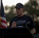 A middle-aged man stands at a podium speaking into a handheld microphone at what appears to be an outdoor commemorative event. He is wearing a black t-shirt referencing the 3rd Infantry Division ('Rock of the Marne, est. 1917') and a black baseball cap. An American flag is visible in the background to his left.