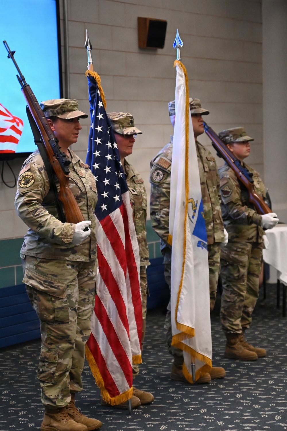 A military color guard ceremony is taking place indoors, with four personnel in OCP (Operational Camouflage Pattern) uniforms presenting flags and carrying ceremonial rifles. The formation includes the United States national flag and what appears to be a light blue Air Force or Space Force organizational flag with gold fringe. The setting appears to be a military dining facility or event hall.