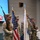 104th Fighter Wing hosts Springfield Chamber of Commerce Breakfast A military color guard ceremony is taking place indoors, with four personnel in OCP (Operational Camouflage Pattern) uniforms presenting flags and carrying ceremonial rifles. The formation includes the United States national flag and what appears to be a light blue Air Force or Space Force organizational flag with gold fringe. The setting appears to be a military dining facility or event hall.