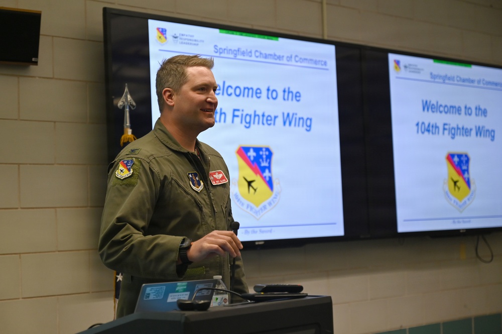 A military officer in an olive green flight suit is presenting at a podium to the Springfield Chamber of Commerce. The presentation screens behind him display 'Welcome to the 104th Fighter Wing' along with the unit's shield insignia. The setting appears to be a briefing room or conference facility at a military installation.