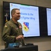 A military officer in an olive green flight suit is presenting at a podium to the Springfield Chamber of Commerce. The presentation screens behind him display 'Welcome to the 104th Fighter Wing' along with the unit's shield insignia. The setting appears to be a briefing room or conference facility at a military installation.