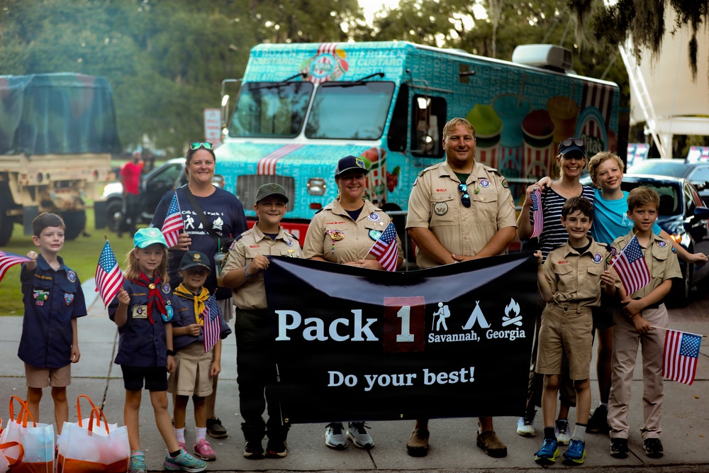 A group of Cub Scouts and adult leaders from Pack 1 in Savannah, Georgia pose together holding a black banner reading 'Pack 1 Savannah, Georgia - Do your best!' Several children and adults are holding small American flags, suggesting a patriotic parade or community event. A colorful teal frozen custard food truck is visible in the background.