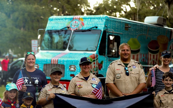 A group of Cub Scouts and adult leaders from Pack 1 in Savannah, Georgia pose together holding a black banner reading 'Pack 1 Savannah, Georgia - Do your best!' Several children and adults are holding small American flags, suggesting a patriotic parade or community event. A colorful teal frozen custard food truck is visible in the background.