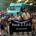A group of Cub Scouts and adult leaders from Pack 1 in Savannah, Georgia pose together holding a black banner reading 'Pack 1 Savannah, Georgia - Do your best!' Several children and adults are holding small American flags, suggesting a patriotic parade or community event. A colorful teal frozen custard food truck is visible in the background.