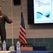 A U.S. Air Force officer in a flight suit is delivering a briefing or presentation in a conference room setting. The presentation slide visible on the large screen compares the F-15 Eagle and F-35 Lightning II aircraft, detailing their respective mission roles. An American flag stands between the presenter and the display screen.
