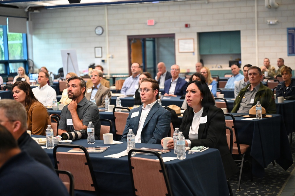 A professional conference or seminar is taking place in a large meeting room, with approximately 30-40 attendees seated at round tables covered with navy blue tablecloths. Participants appear to be attentively watching a presentation or speaker located off-camera to the front. The setting suggests a formal business or organizational event, with attendees wearing business casual to business professional attire.