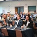 A professional conference or seminar is taking place in a large meeting room, with approximately 30-40 attendees seated at round tables covered with navy blue tablecloths. Participants appear to be attentively watching a presentation or speaker located off-camera to the front. The setting suggests a formal business or organizational event, with attendees wearing business casual to business professional attire.