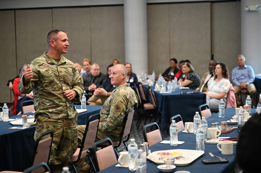 A military officer in OCP (Operational Camouflage Pattern) uniform is standing and addressing an audience in what appears to be a conference or banquet hall setting. The speaker's name tape reads 'GARDNER' and he is gesturing while speaking to a mixed audience of military personnel and civilians seated at round tables. A second uniformed military member is seated nearby listening to the presentation.
