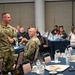 A military officer in OCP (Operational Camouflage Pattern) uniform is standing and addressing an audience in what appears to be a conference or banquet hall setting. The speaker's name tape reads 'GARDNER' and he is gesturing while speaking to a mixed audience of military personnel and civilians seated at round tables. A second uniformed military member is seated nearby listening to the presentation.