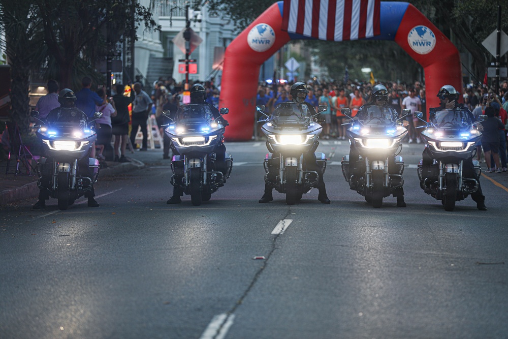 Five police motorcycles with headlights on are lined up on a street in formation, appearing to lead or escort a public event. Behind them is a red inflatable arch branded with 'MWR' and decorated with an American flag, suggesting a military or community event. Large crowds of spectators line both sides of the street.