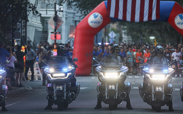 Five police motorcycles with headlights on are lined up on a street in formation, appearing to lead or escort a public event. Behind them is a red inflatable arch branded with 'MWR' and decorated with an American flag, suggesting a military or community event. Large crowds of spectators line both sides of the street.