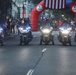 Five police motorcycles with headlights on are lined up on a street in formation, appearing to lead or escort a public event. Behind them is a red inflatable arch branded with 'MWR' and decorated with an American flag, suggesting a military or community event. Large crowds of spectators line both sides of the street.