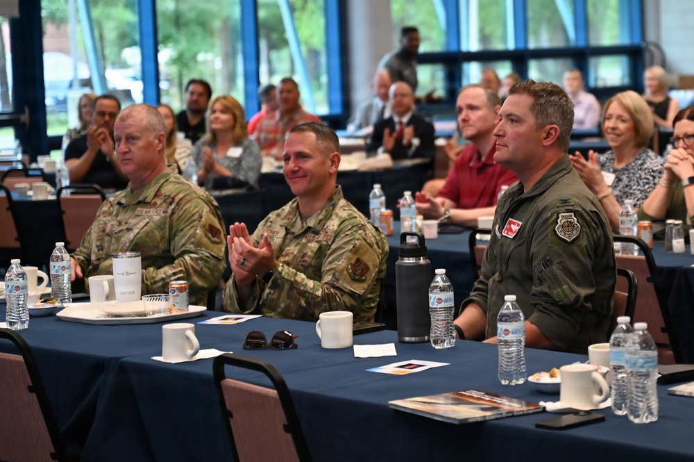 A formal military and civilian gathering, likely a briefing, luncheon, or awards ceremony, taking place in a conference or banquet hall. Three military personnel in the foreground — two in Army Combat Uniform (OCP pattern) and one in an olive green flight suit — are seated at a table with refreshments. The audience behind them appears to be applauding, suggesting a presentation or recognition moment is occurring.