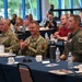 A formal military and civilian gathering, likely a briefing, luncheon, or awards ceremony, taking place in a conference or banquet hall. Three military personnel in the foreground — two in Army Combat Uniform (OCP pattern) and one in an olive green flight suit — are seated at a table with refreshments. The audience behind them appears to be applauding, suggesting a presentation or recognition moment is occurring.
