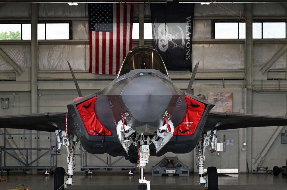 A front-facing view of an F-35 Lightning II stealth fighter jet parked inside a military hangar. The aircraft has its weapon bay doors open, revealing internal bay mechanisms, and is fitted with red 'Remove Before Flight' safety covers. An American flag and a black 'WEAPONS' squadron banner are visible in the background.