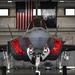 A front-facing view of an F-35 Lightning II stealth fighter jet parked inside a military hangar. The aircraft has its weapon bay doors open, revealing internal bay mechanisms, and is fitted with red 'Remove Before Flight' safety covers. An American flag and a black 'WEAPONS' squadron banner are visible in the background.
