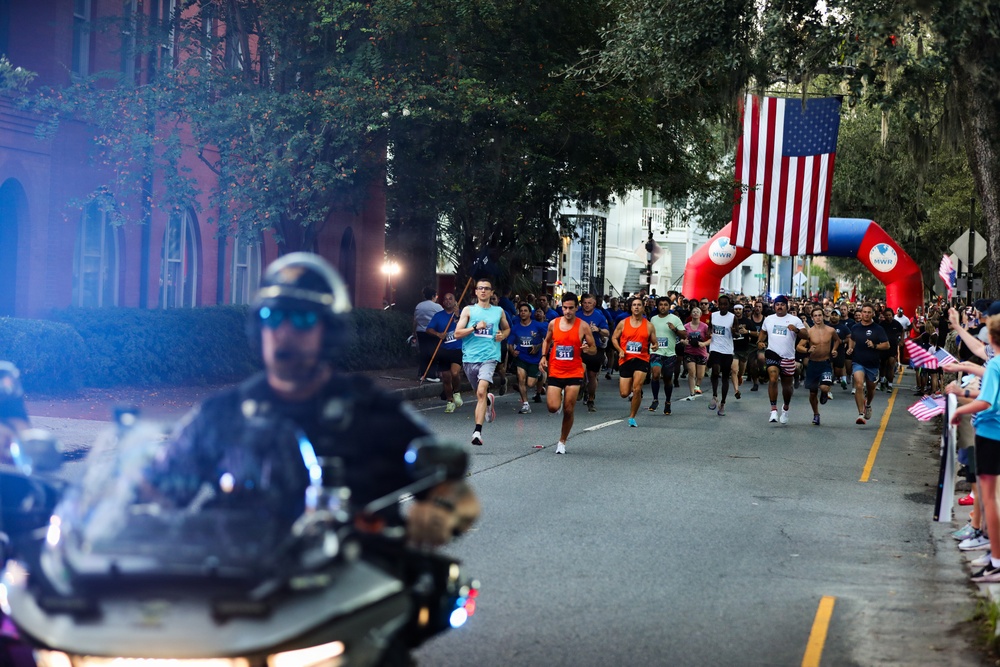 A road race is underway on a tree-lined urban street, with a large crowd of runners departing from a red and blue inflatable arch start/finish gate bearing 'MWR' branding. A large American flag is suspended above the arch, and spectators line the right side of the road waving small American flags, suggesting a patriotic or military-affiliated event. A police motorcycle officer leads the pack of runners down the course.