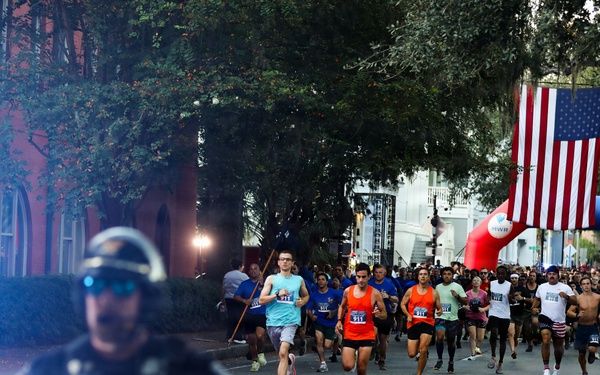 A road race is underway on a tree-lined urban street, with a large crowd of runners departing from a red and blue inflatable arch start/finish gate bearing 'MWR' branding. A large American flag is suspended above the arch, and spectators line the right side of the road waving small American flags, suggesting a patriotic or military-affiliated event. A police motorcycle officer leads the pack of runners down the course.