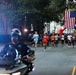 A road race is underway on a tree-lined urban street, with a large crowd of runners departing from a red and blue inflatable arch start/finish gate bearing 'MWR' branding. A large American flag is suspended above the arch, and spectators line the right side of the road waving small American flags, suggesting a patriotic or military-affiliated event. A police motorcycle officer leads the pack of runners down the course.