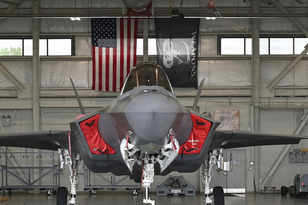 A front-facing view of an F-35 Lightning II stealth fighter jet parked inside a military hangar. The aircraft has its weapon bay doors open, revealing red safety covers and 'Remove Before Flight' tags. An American flag and a black 'WEAPONS' banner are prominently displayed on the hangar wall behind the aircraft.